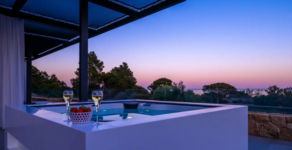Un jacuzzi de l'Hôtel Villa Padulaccia au crépuscule sous une pergola, deux flûtes et une coupelle de fraises sur le rebord, la mer au loin