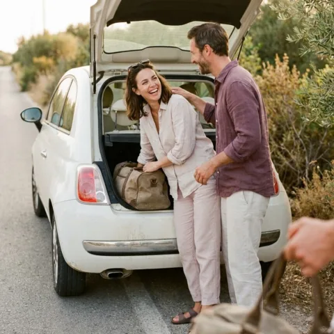 Un couple rit à l'arrière d'une petite voiture blanche, coffre ouvert, en chargeant un sac de voyage au bord d'une route de campagne