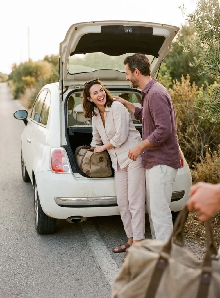 Un couple rit à l'arrière d'une petite voiture blanche, coffre ouvert, en chargeant un sac de voyage au bord d'une route de campagne