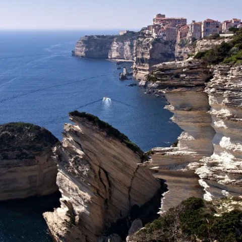 La ville haute de Bonifacio au bord des falaises blanches, vue depuis le site protégé de Campo Romanello, avec le rocher du Grain de Sable au premier plan et deux bateaux en mer.