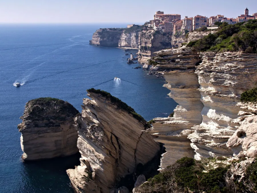 La ville haute de Bonifacio au bord des falaises blanches, vue depuis le site protégé de Campo Romanello, avec le rocher du Grain de Sable au premier plan et deux bateaux en mer.