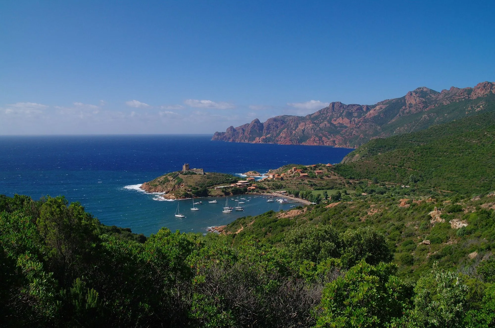 La baie de Girolata et son fortin génois, voiliers au mouillage, falaises rouges du golfe en arrière-plan.