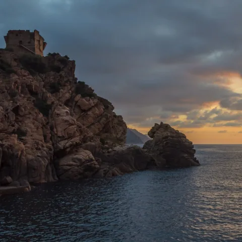 La tour génoise de Porto sur son rocher de granit, au soir, sous des nuages sombres, au-dessus du golfe de Porto.
