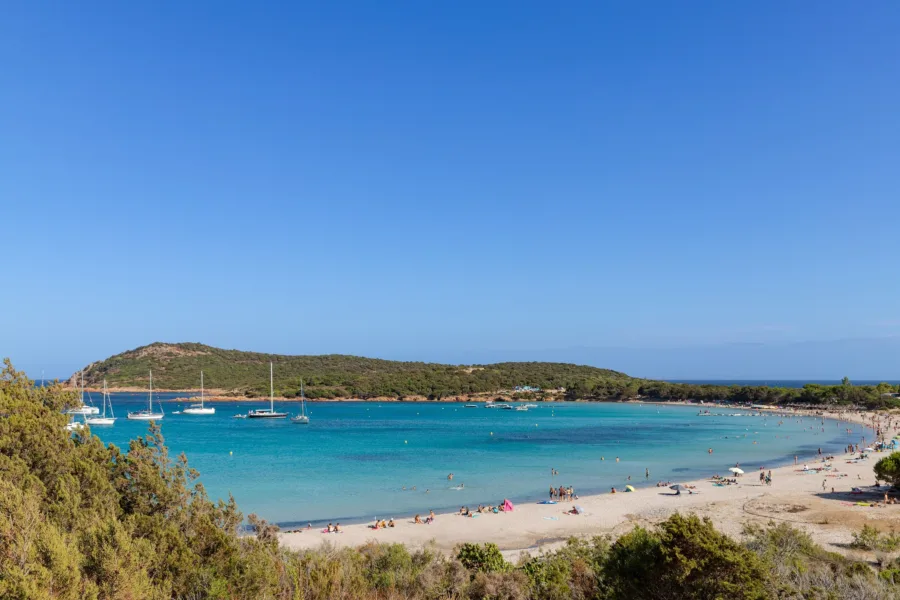 La baie en fer à cheval de Rondinara vue depuis le maquis, eau turquoise, voiliers au mouillage et plage fréquentée (juillet 2022).
