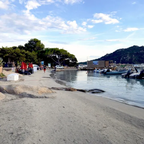 Fin de saison sur la plage de Santa Giulia : sable presque vide, bateaux amarrés au ponton, collines en fond.